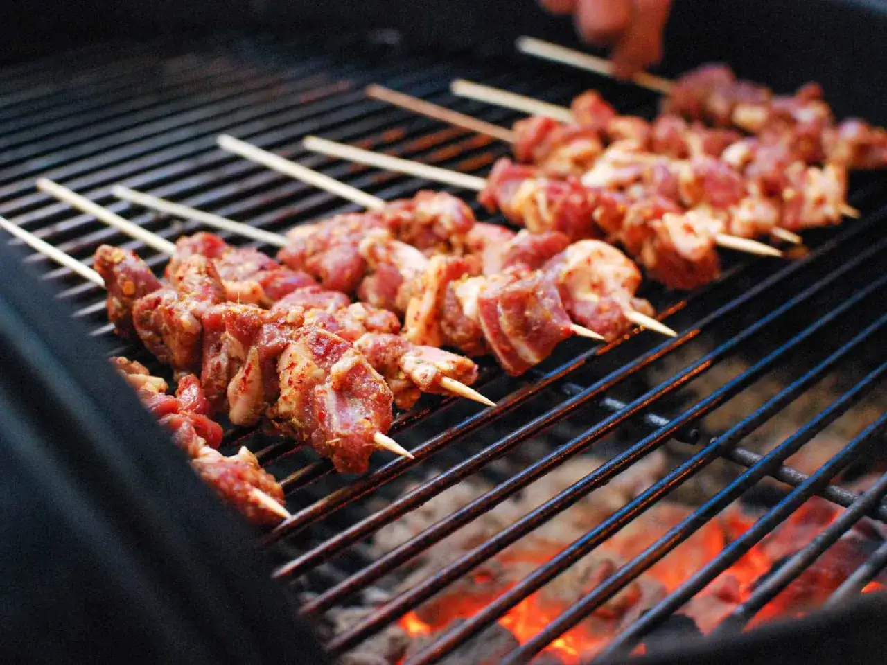 Plusieurs brochettes d'agneau crues, assaisonnées, cuisent sur un gril chaud avec des braises rougeoyantes en dessous.
