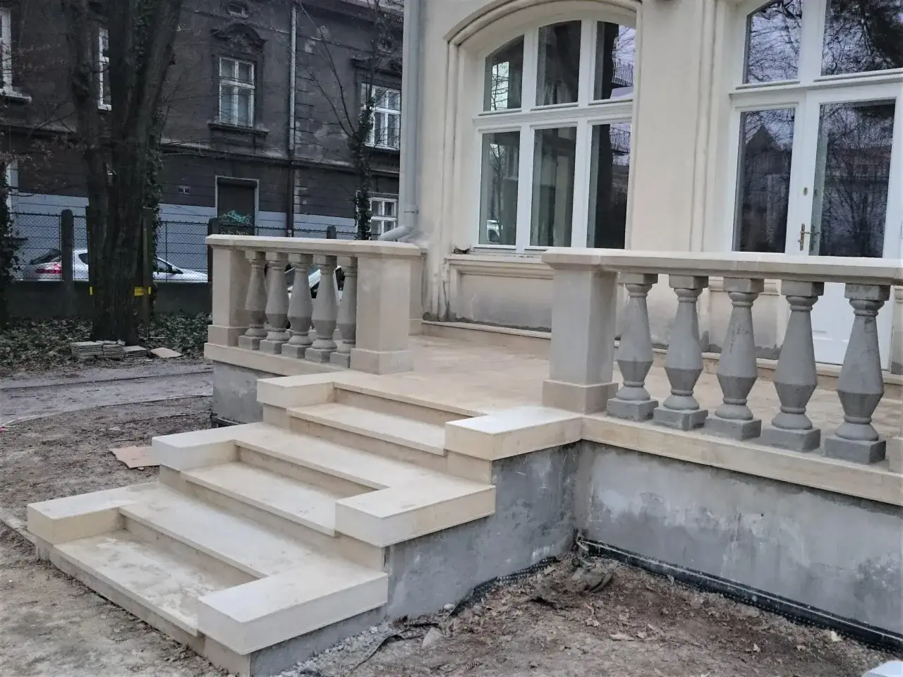 Schody i balustrada z piaskowca przed budynkiem. W tle widać starą kamienicę i drzewa.