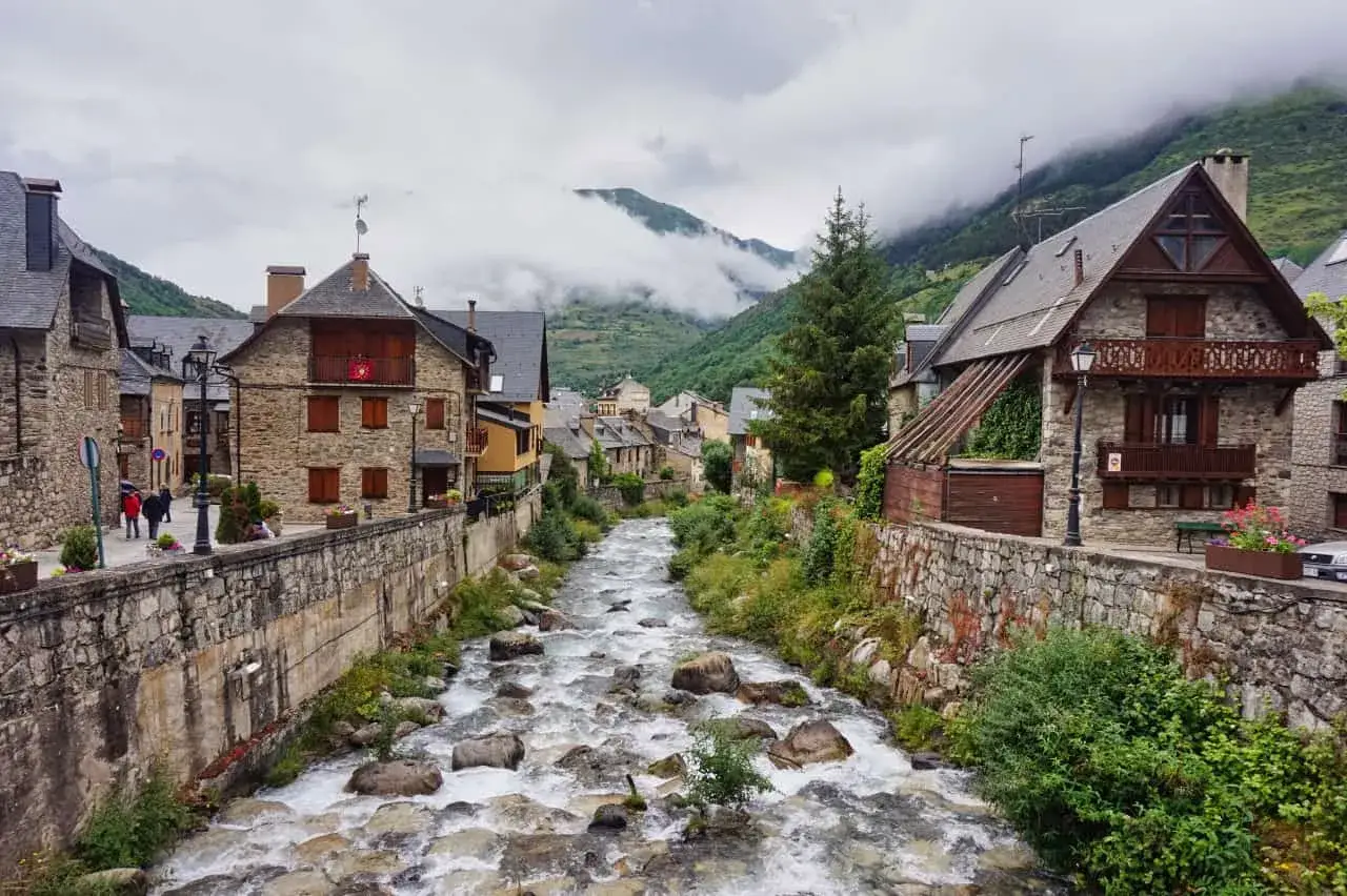 Ein Fluss schl&auml;ngelt sich durch das malerische Val d'Aran, ges&auml;umt von traditionellen Steinh&auml;usern und gr&uuml;nen Bergen im Nebel.