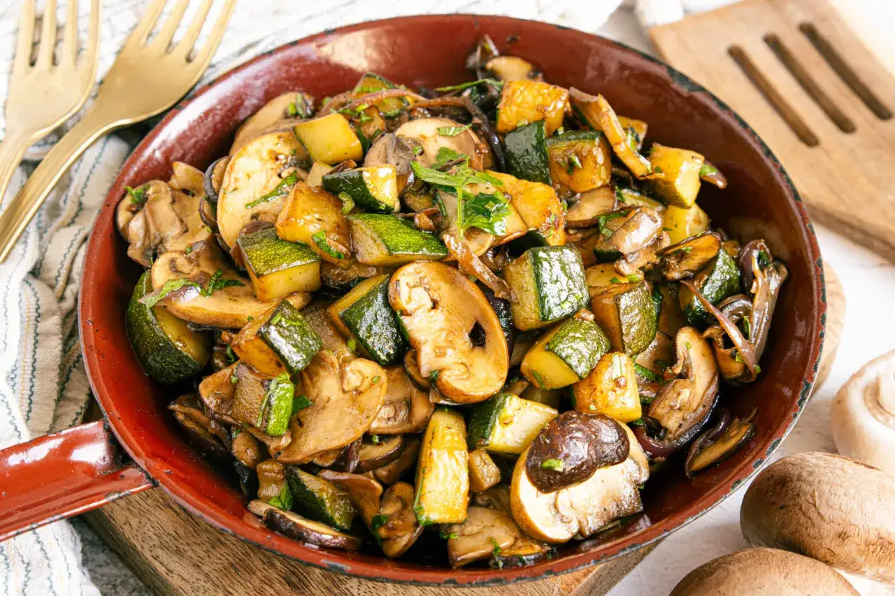 Herzhafte zucchini pilz pfanne, mit Kräutern verfeinert, dampft in einer roten Pfanne. Goldene Gabeln liegen bereit.