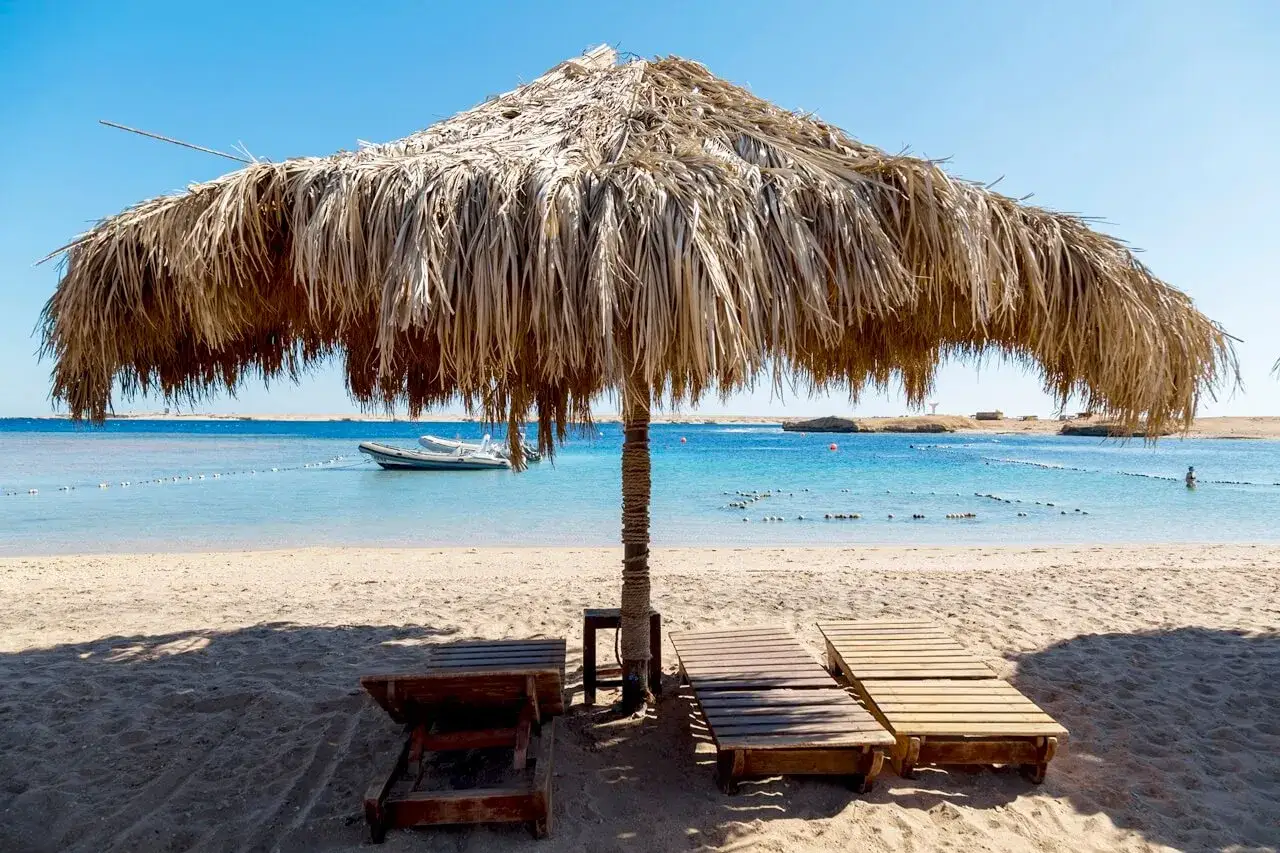 Słomiany parasol chroni leżaki na piaszczystej plaży. W tle błękitne morze, ł&oacute;dź i linia brzegowa. Idealne miejsce, kiedy do Egiptu na wakacje.