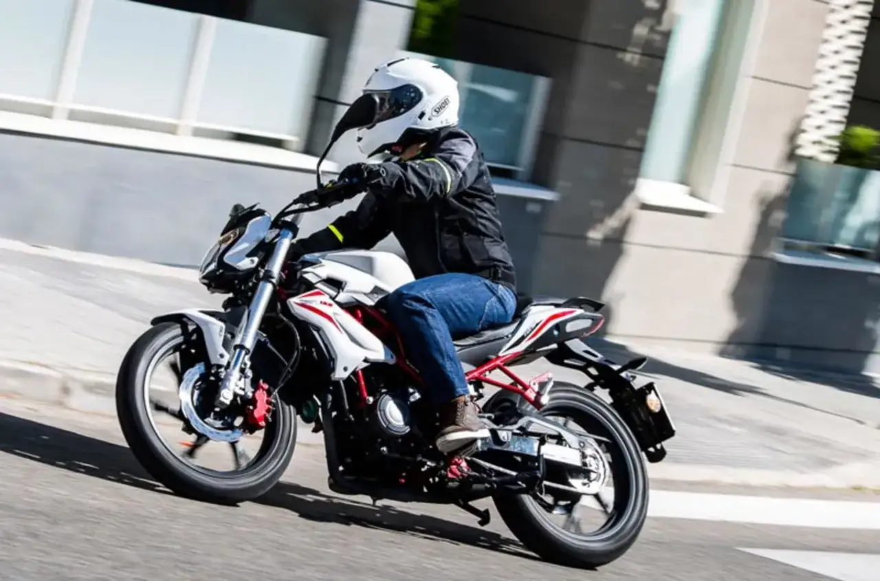 Motociclista en una Benelli BN 125 blanca y roja, disfrutando de un paseo urbano. Las opiniones sobre la Benelli BN 125 la describen como &aacute;gil y divertida.