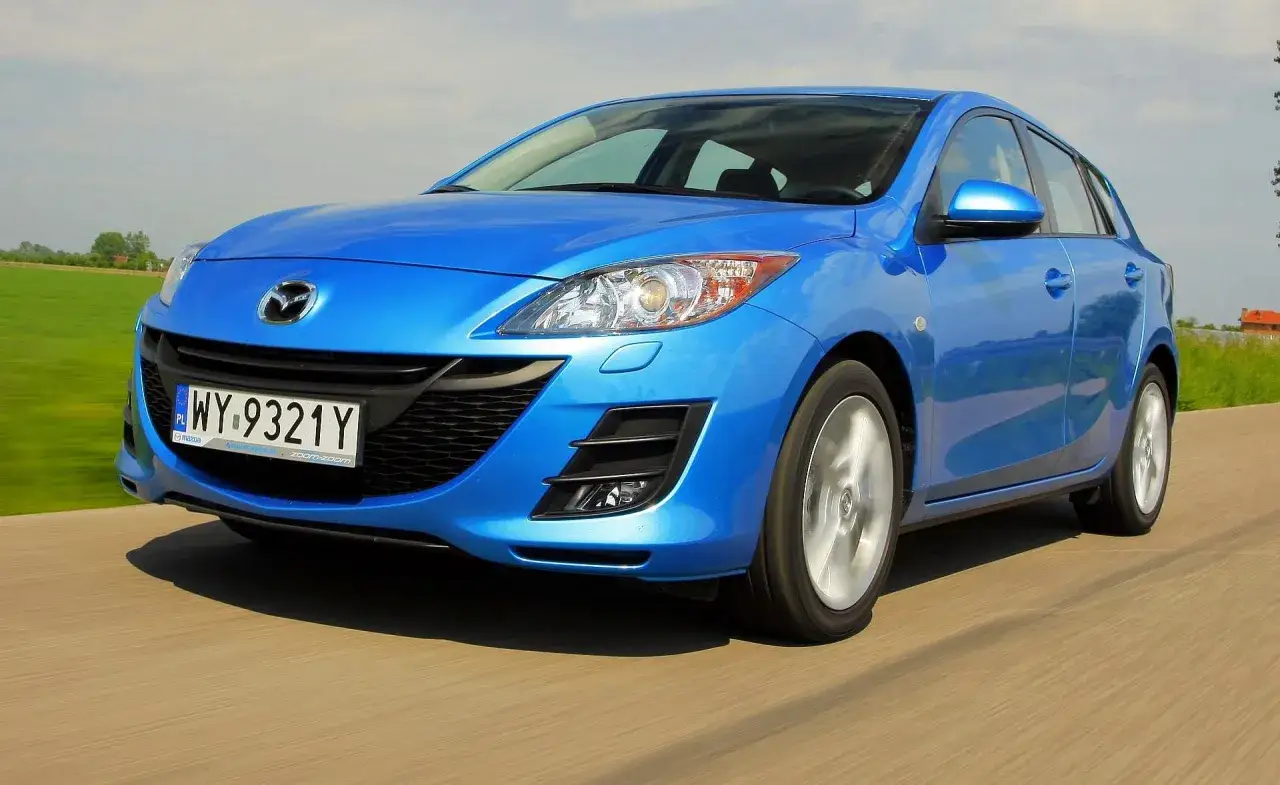 Niebieska Mazda 3 2010 pędzi po drodze, za oknem zielone pola i błękitne niebo.