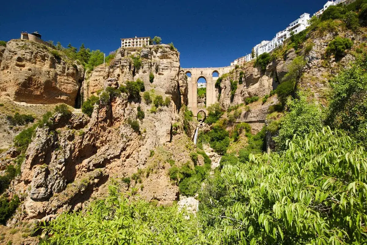 A dramatic **Ronda bridge viewpoint** showcasing the Puente Nuevo spanning a deep gorge, with buildings perched on cliffs and a waterfall cascading below.