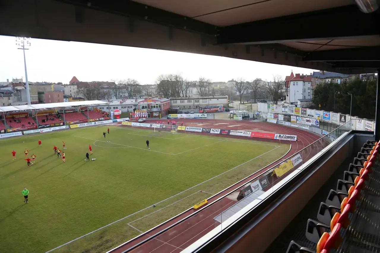 Piłkarze trenują na murawie stadionu miejskiego w Chojnicach. W tle trybuny i budynki.