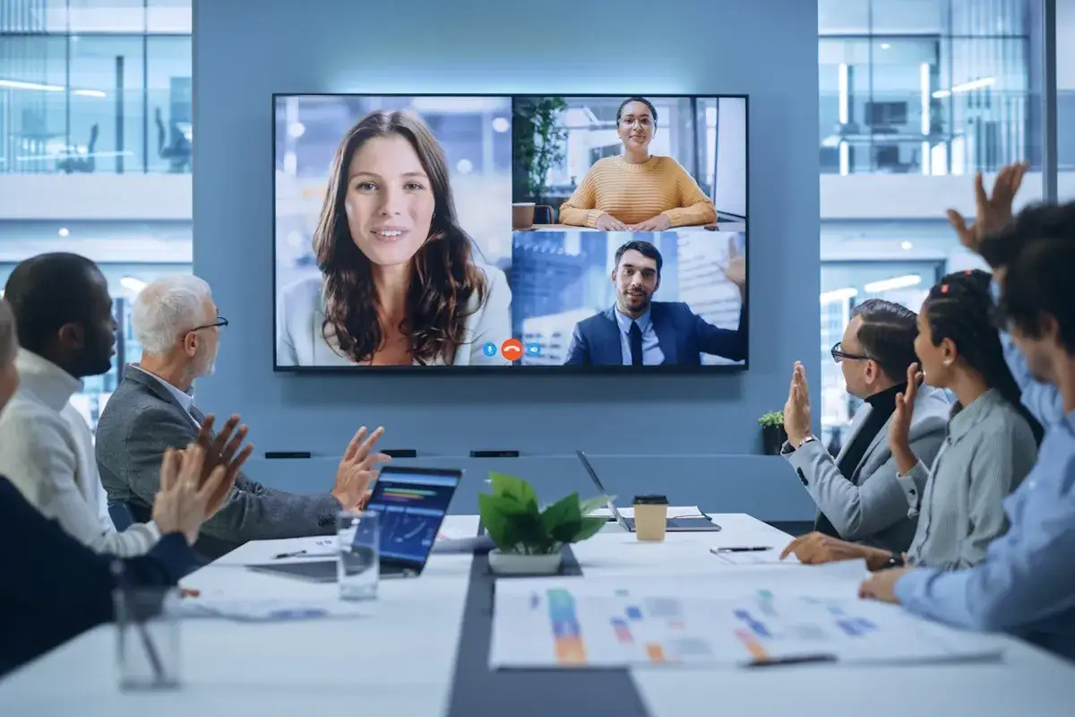 A team in a modern office engages in a video conference, fostering communication and accountability. They are actively participating, with some raising hands, ensuring everyone is heard and understood.