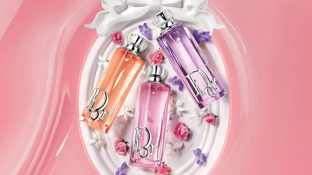 Trzy butelki perfum Dior Addict w odcieniach różu, pomarańczy i fioletu, otoczone delikatnymi kwiatami i satynową wstęgą.