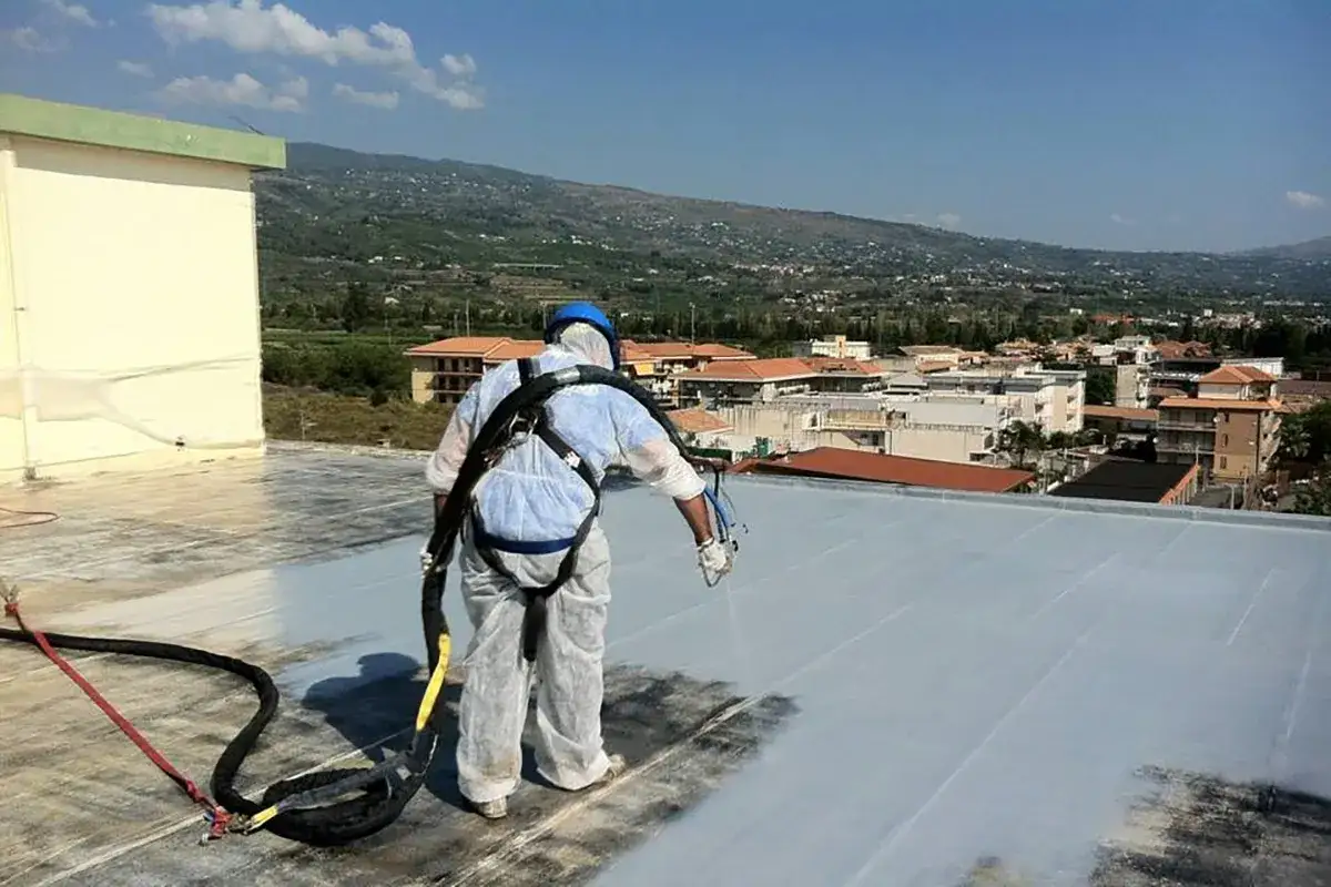 Operaio in tuta protettiva esegue impermeabilizzazione tetti a spruzzo su un edificio, con vista panoramica su un paese collinare.