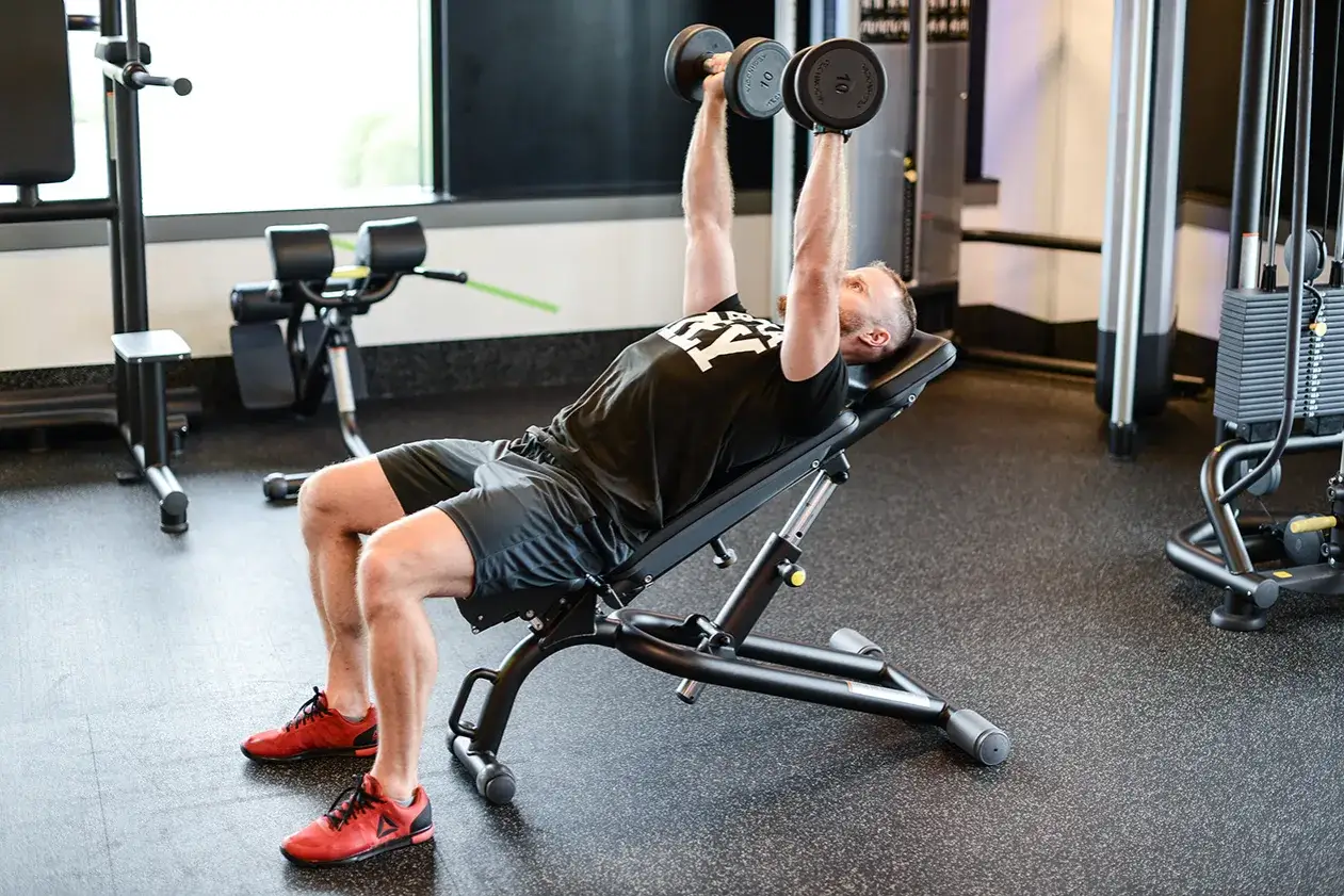 Mężczyzna wykonuje ćwiczenie incline press z hantlami na ławce skośnej w siłowni.