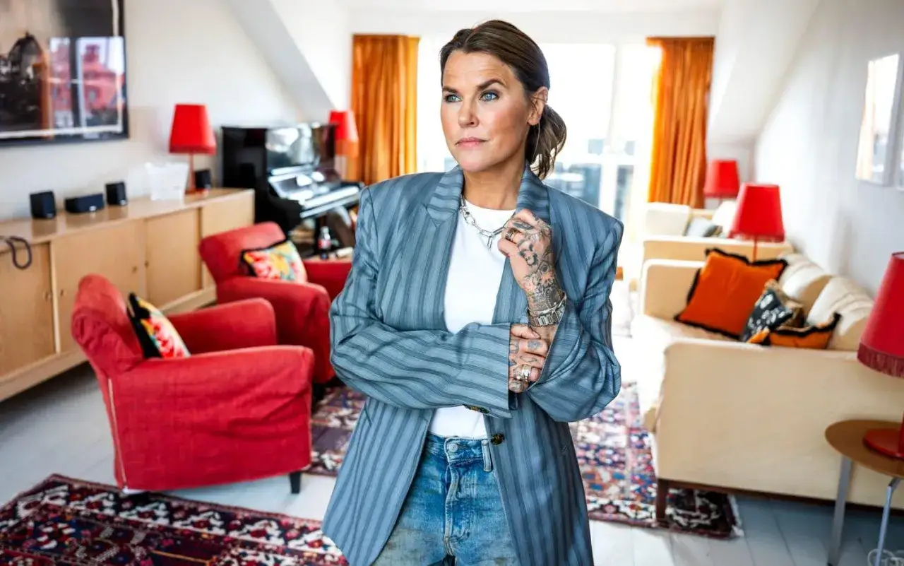 En kvinna med tatueringar står i ett vardagsrum med röda fåtöljer och en soffa. Hon bär en randig kavaj och jeans. Kanske hon funderar på vilka bra serier hon ska se ikväll.