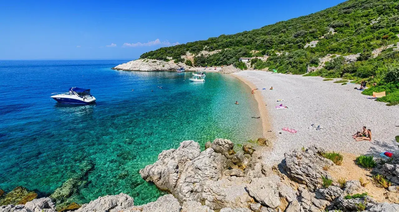 Idealne miejsce na wakacje w Chorwacji: kamienista plaża, turkusowa woda, łodzie i zielone wzgórza.