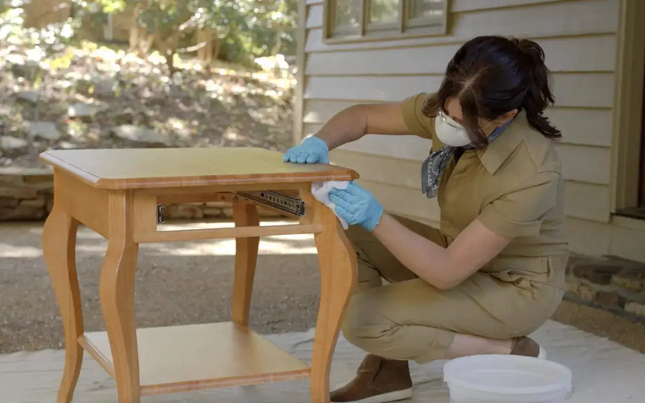 Préparation d'une table en bois pour la peinture. Une femme, portant masque et gants, nettoie la surface.