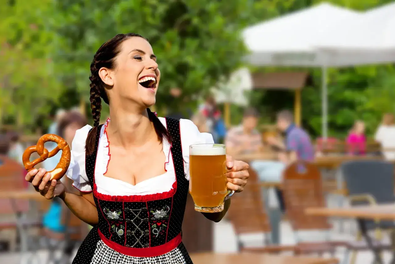 Uśmiechnięta kobieta w stroju bawarskim trzymająca kufel piwa i precel na tle ogródka piwnego podczas Oktoberfestu w Monachium.