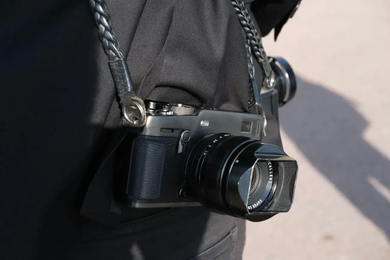 En person b&auml;r en svart jacka med en fl&auml;tad l&auml;derrem som h&aring;ller en Fuji Xpro 3-kamera.
