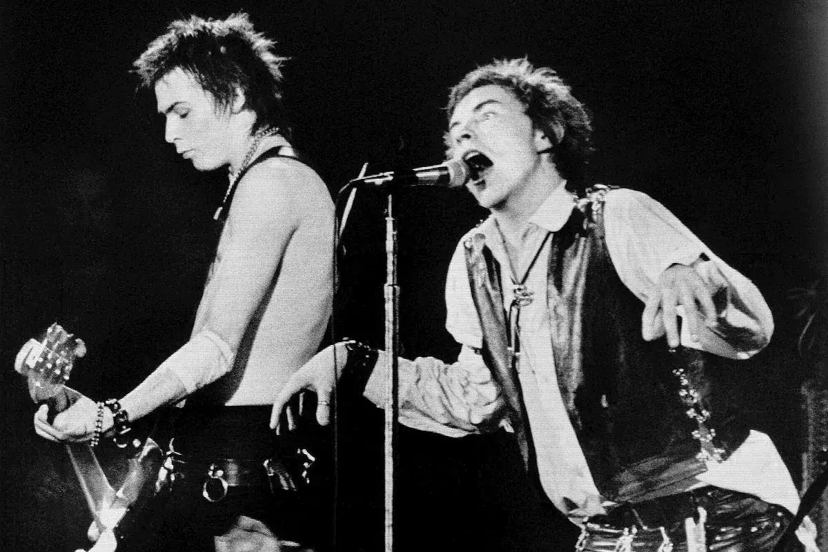 Sid Vicious z zespołu Sex Pistols gra na gitarze, podczas gdy wokalista śpiewa do mikrofonu.