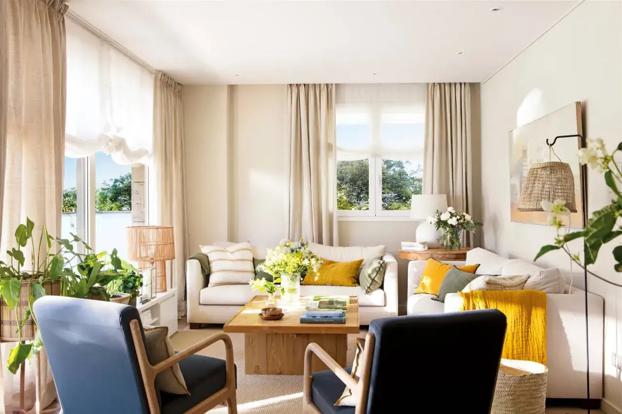 Estor de lino y cortinas de tela vaporosa en un salón luminoso con sofás blancos, sillones azules y mesa de centro de madera.