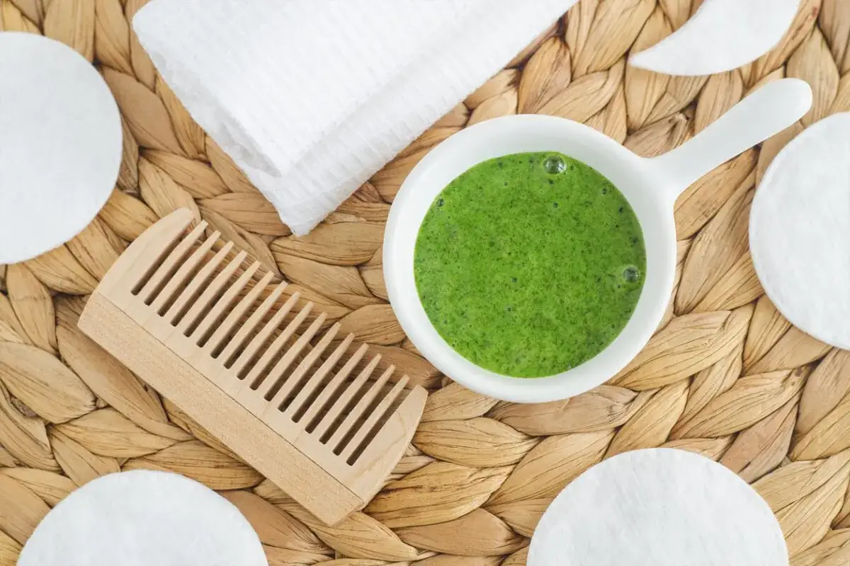 Un tazón con una mascarilla de te verde para el pelo, junto a un peine de madera, toalla y discos de algodón.