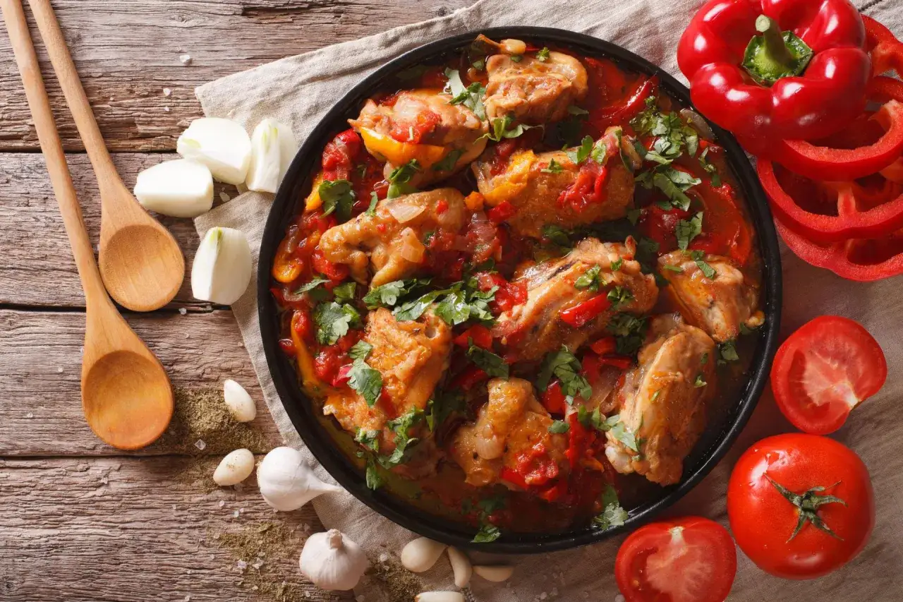 Un plat de poulet basquaise mijot&eacute; avec des poivrons rouges, des tomates et des oignons, garni de persil frais. Des ingr&eacute;dients frais et des cuill&egrave;res en bois compl&egrave;tent la sc&egrave;ne.