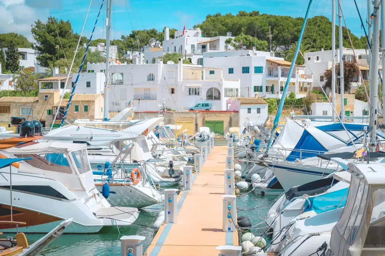 Boote im Hafen von Cala d'Or Altstadt, wei&szlig;e H&auml;user mit roten Ziegeld&auml;chern im Hintergrund, sonniger Tag.