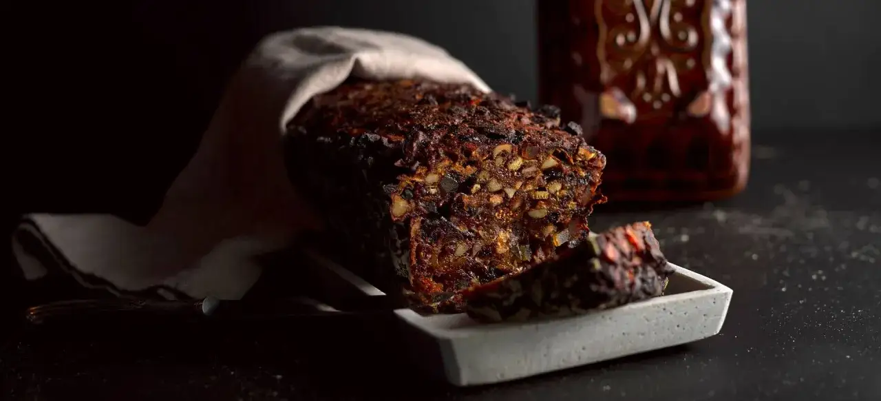 En m&ouml;rk, saftig sockerfri t&aring;rta med n&ouml;tter och torkad frukt, delvis t&auml;ckt av en duk. En bit ligger p&aring; en gr&aring; bricka.