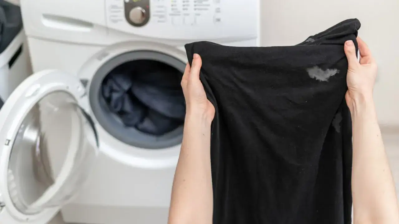 Manos sostienen ropa negra con manchas blancas, junto a una lavadora abierta. &iquest;Por qu&eacute; la ropa negra sale con manchas blancas?