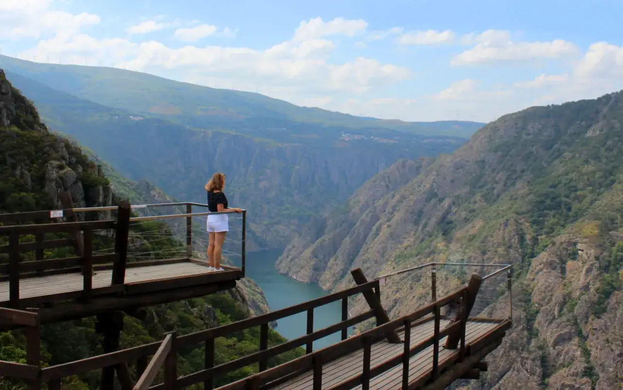 Mujer contemplando la inmensidad de los cañones del Sil desde una pasarela de madera. El río serpentea entre montañas escarpadas bajo un cielo azul con nubes.