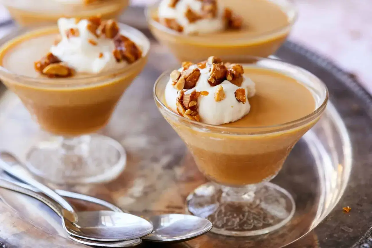 Trois coupes de panna cotta caramel, garnies de cr&egrave;me fouett&eacute;e et de noix caram&eacute;lis&eacute;es, sur un plateau argent&eacute; avec des cuill&egrave;res.