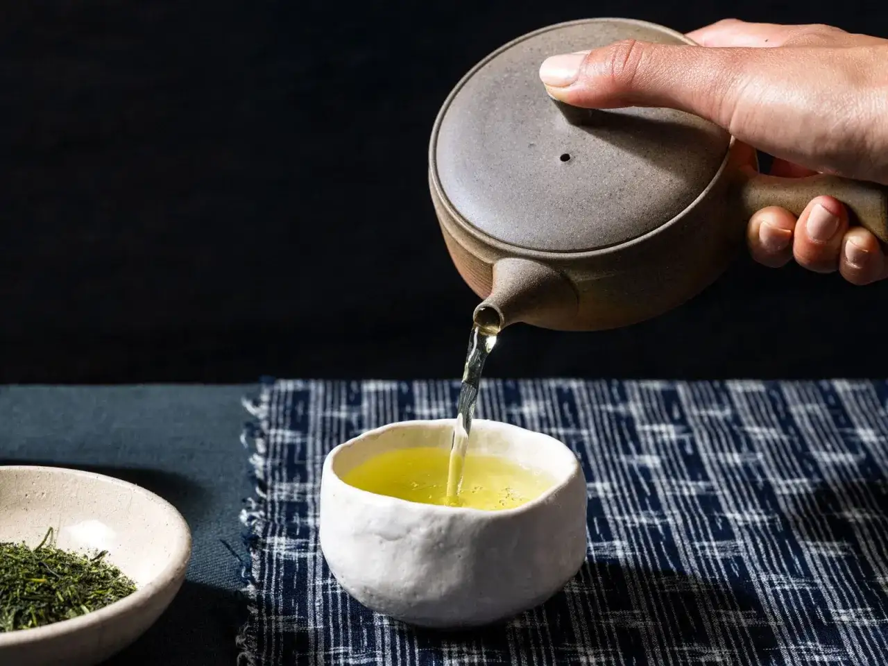 Dłoń nalewa zieloną herbatę sencha z glinianego czajniczka do ceramicznej filiżanki. Obok miseczka z liśćmi herbaty.
