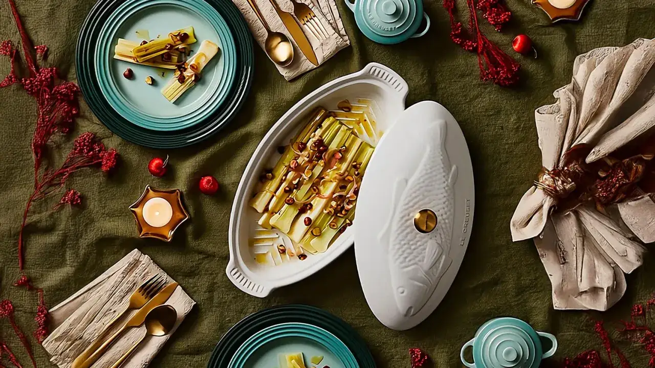 Puerros al horno, tiernos y dorados, servidos en una elegante fuente con tapa en forma de pez. Decoración festiva con velas y frutos rojos.