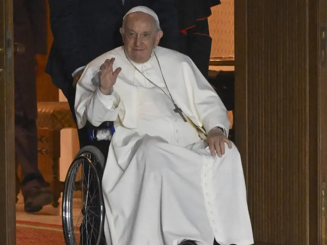 Papież Franciszek w białej szacie, siedząc na wózku inwalidzkim, macha do wiernych. Wiek papieża nie jest widoczny na zdjęciu.