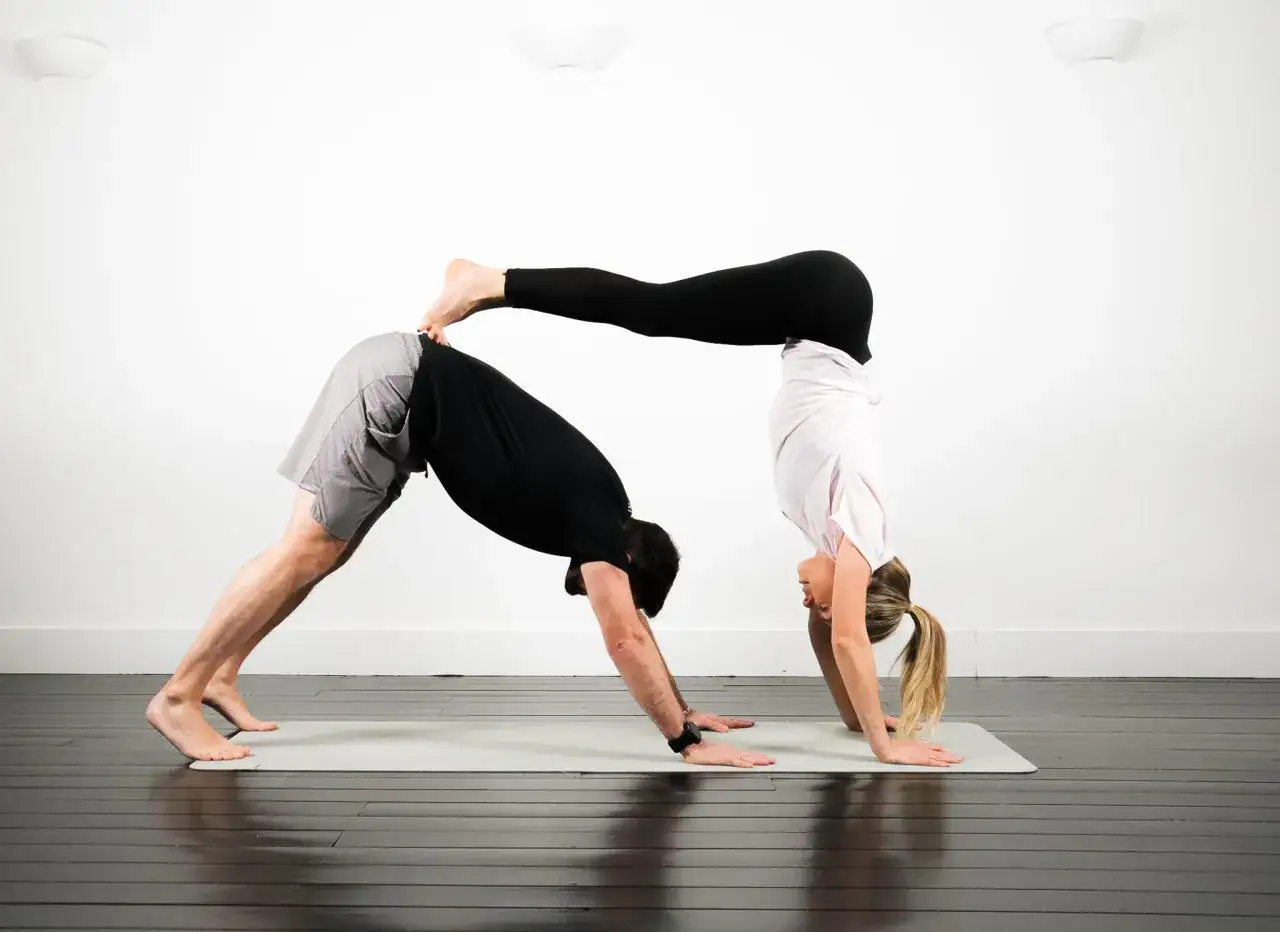 Två personer utför avancerad yoga. Mannen är i nedåtgående hund, kvinnan balanserar på hans rygg i en handstående.