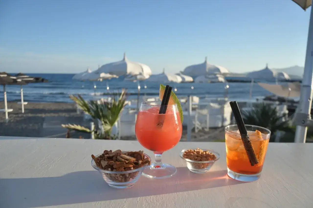 Aperitivo riccione: due cocktail colorati e ciotole di snack sulla spiaggia al tramonto.