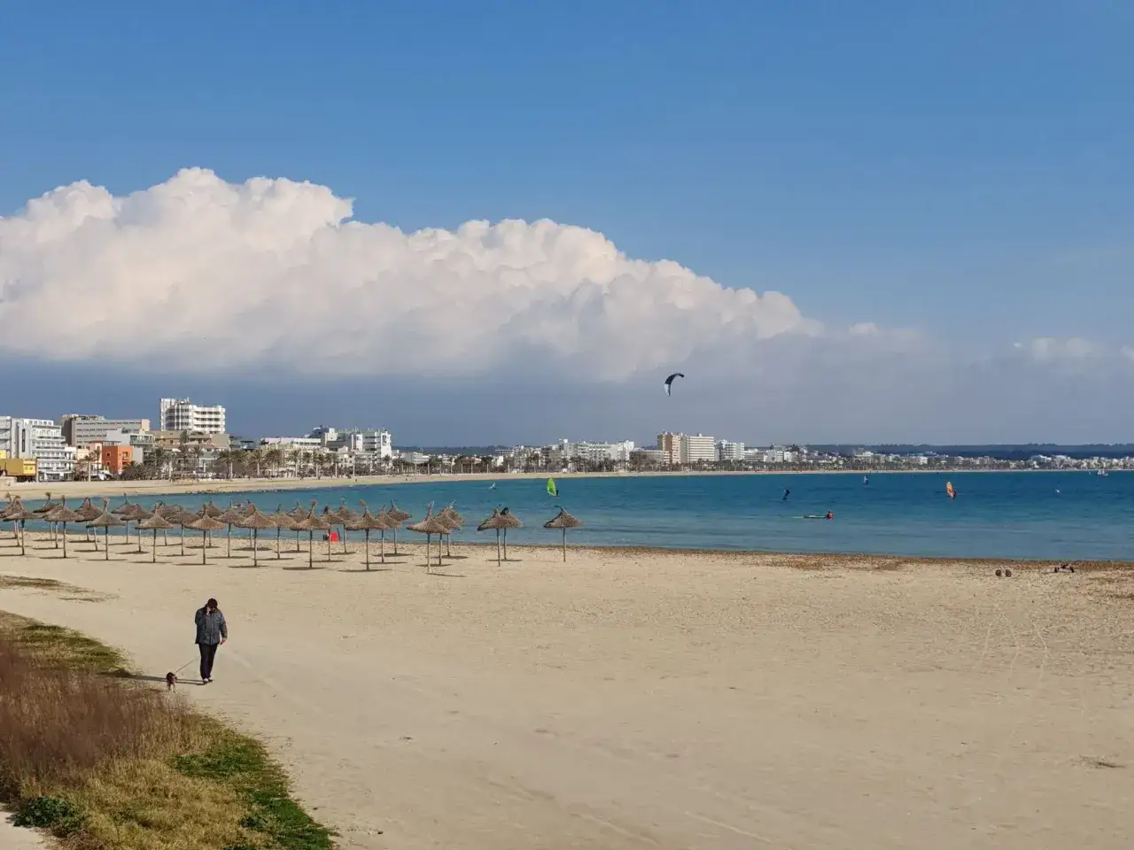 Spacerem po plaży w Can Pastilla, z widokiem na morze, parasole i miasto. Na wodzie kitesurferzy.