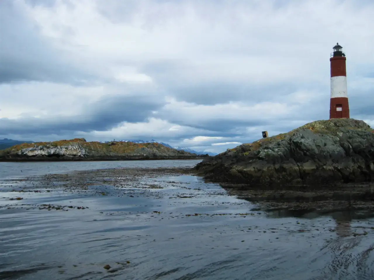 Latarnia morska na skalistej wysepce, symbol krajobrazu **Tierra del Fuego**. Woda i chmury tworzą surowy, północny klimat.