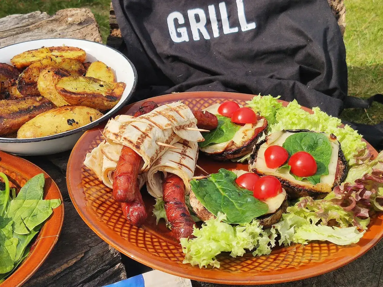 Pyszne dodatki do grilla: kiełbaski zawinięte w tortillę, pieczone ziemniaki i grillowane pieczarki z pomidorkami.