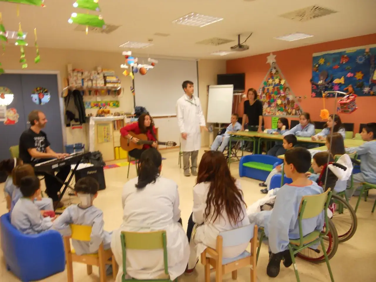 Un grupo de ni&ntilde;os y adultos en un aula, algunos tocando m&uacute;sica. Un hombre toca el teclado, una mujer toca la guitarra. Esto es como ser un buen profesor, involucrando a todos.