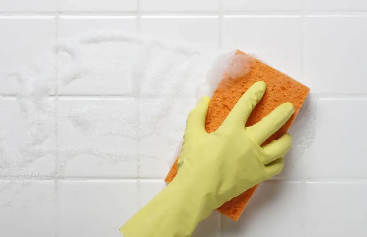 A gloved hand scrubs white shower tiles with a soapy orange sponge, demonstrating how to clean shower tiles effectively.