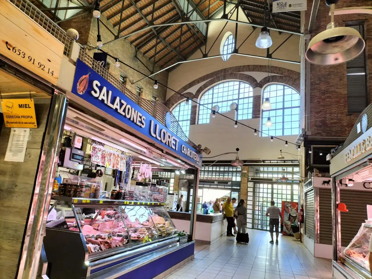 Interior del mercat central d'Alacant, con puestos de charcutería y salazones, y gente comprando.