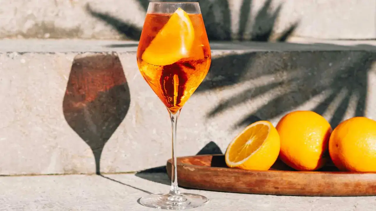Un verre de spritz v&eacute;nitien, garni d'une tranche d'orange, avec des oranges sur un plateau en bois.