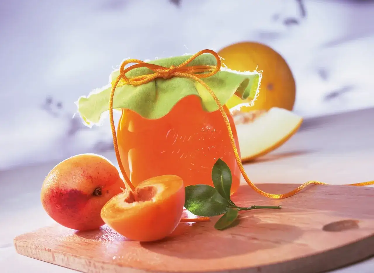 Ein Glas leuchtend orangefarbener Melonen Marmelade, verziert mit einem grünen Tuch und einer Schnur. Daneben liegen frische Aprikosen und ein Stück Melone.