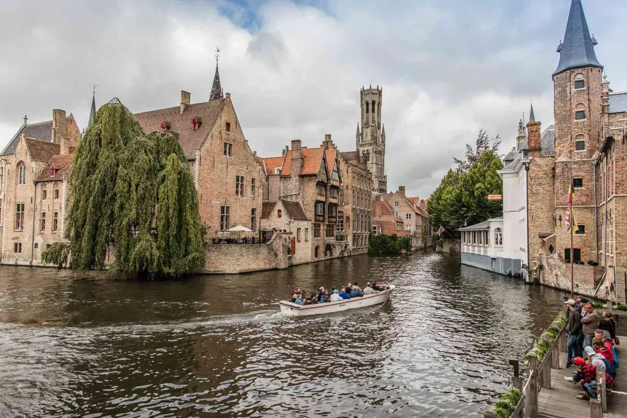 En båt med turister glider fram på en kanal i Brygge, Belgien, omgiven av historiska byggnader och ett högt klocktorn.
