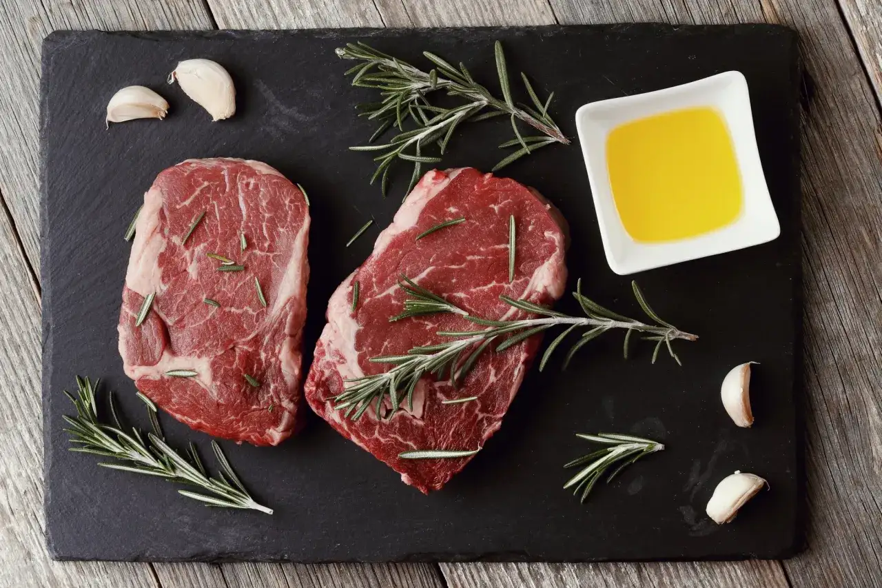 Dwa surowe steki wołowe z rozmarynem, czosnkiem i oliwą. Idealne składniki na pyszne bbq. Co to znaczy? To zapowiedź uczty!