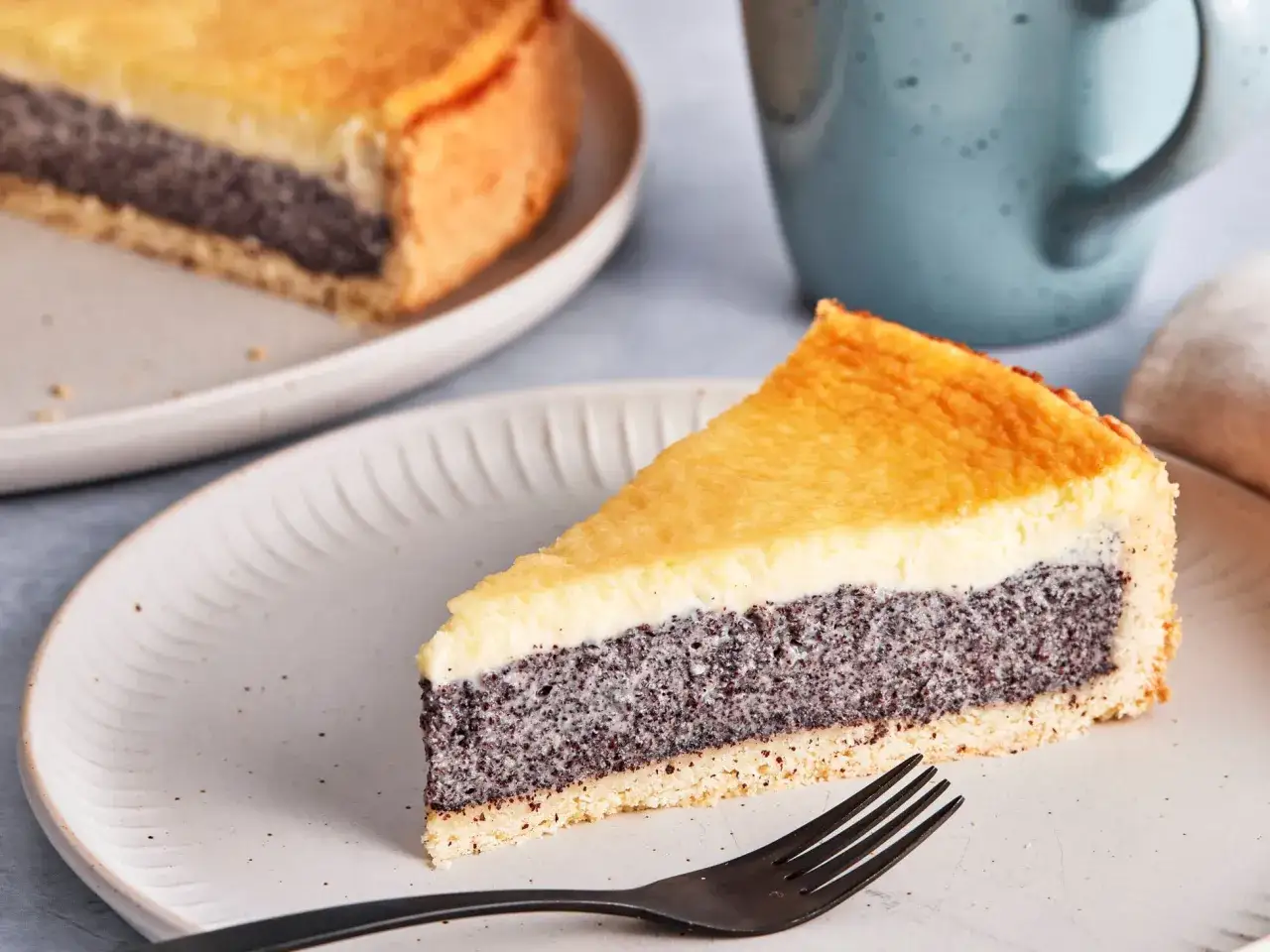 Ein Stück Mohn Schmand Kuchen auf einem Teller, mit einer Gabel daneben. Der Kuchen hat eine goldbraune Kruste und eine cremige Füllung.