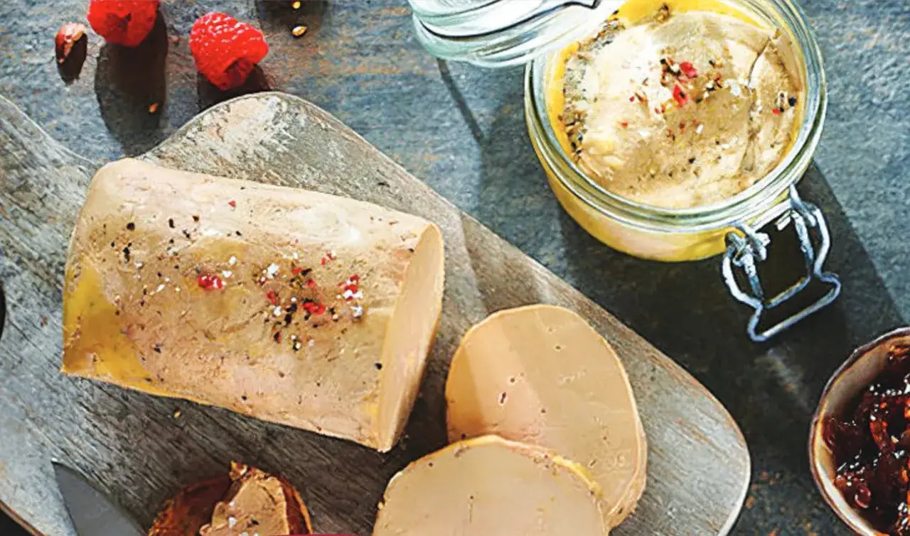 Un bloc de foie gras de qualité, assaisonné de poivre et de baies roses, est présenté sur une planche en bois avec des framboises et une petite coupelle de confiture d'oignons.
