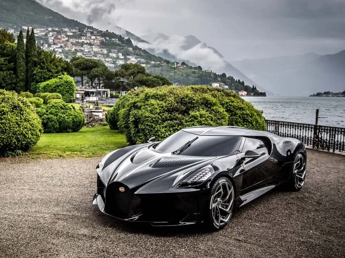 Un elegante Bugatti negro, el coche m&aacute;s caro del mundo, descansa junto a un lago sereno, con un pueblo pintoresco en la ladera de una monta&ntilde;a.