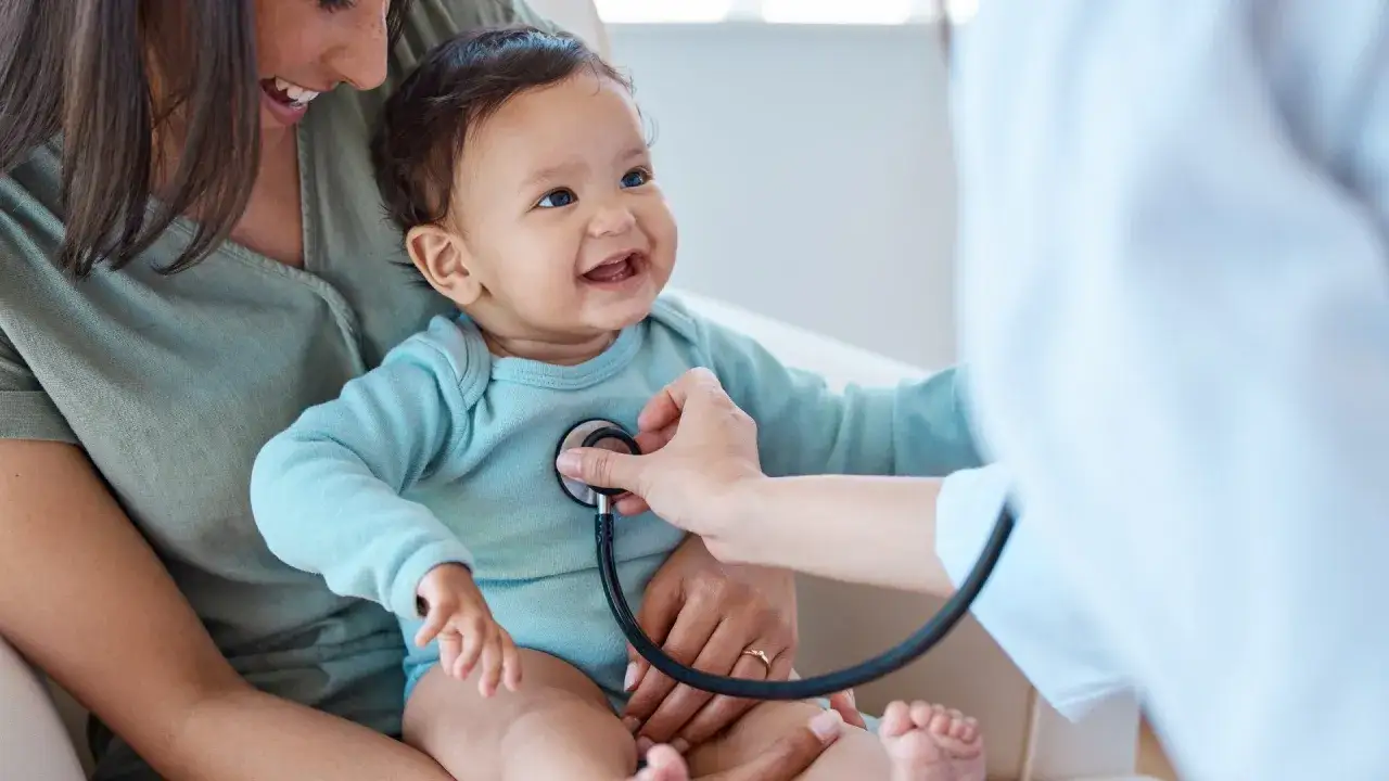 Un neonato di 2 mesi sorride mentre il medico ascolta il suo petto con uno stetoscopio. La mamma lo tiene in braccio.