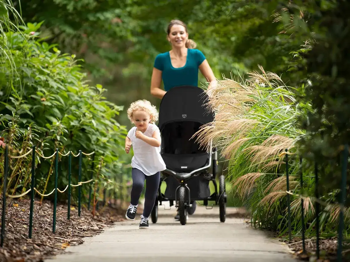 Eine Frau joggt mit Kinderwagen durch einen Park, w&auml;hrend ihr kleines Kind vor ihr rennt.