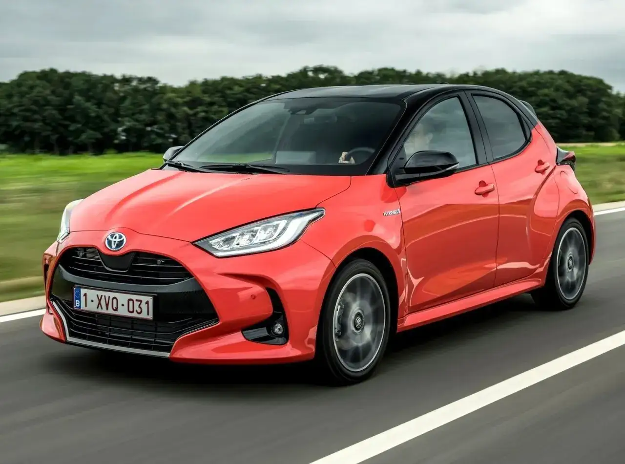 Czerwony Toyota Yaris IV z czarnym dachem pędzi po drodze, otoczony zielenią.