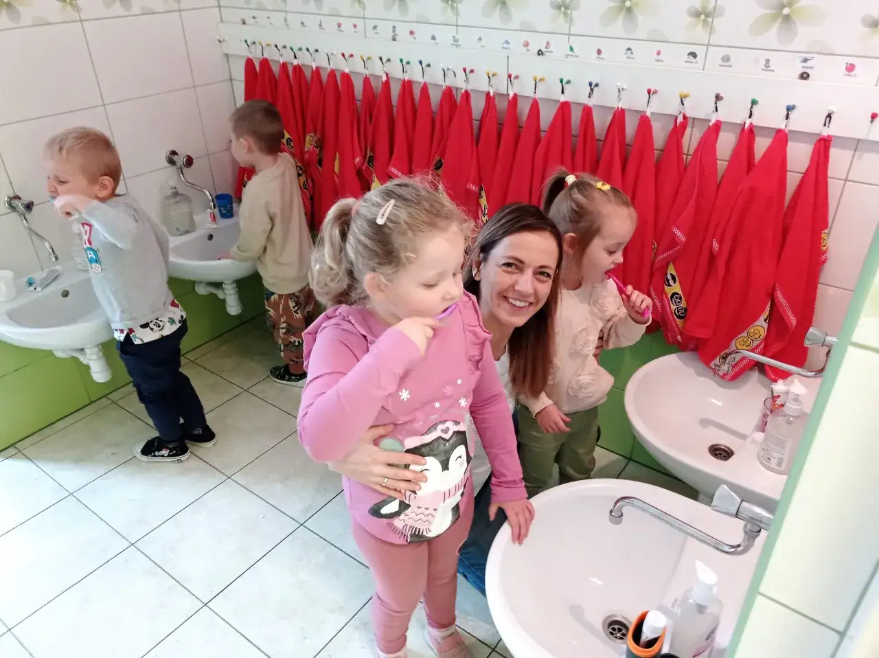 Uśmiechnięta pani pokazuje dzieciom, jak nauczyć dziecko myć zęby. Maluchy szczotkują ząbki przy umywalkach, a nad nimi wiszą czerwone ręczniki.