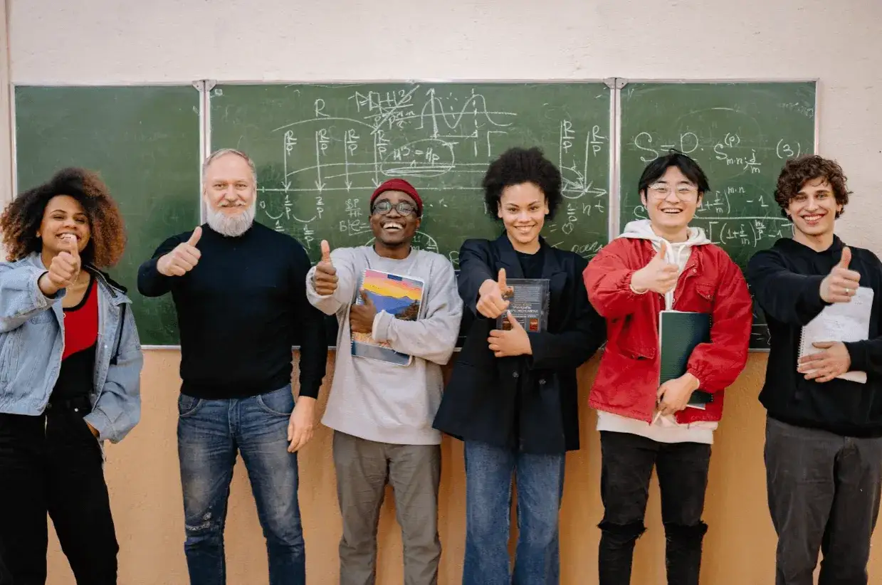 Un grupo diverso de estudiantes y un profesor sonríen y dan pulgares arriba frente a un pizarrón con ecuaciones. Un ejemplo de intercambio cultural en el aula.